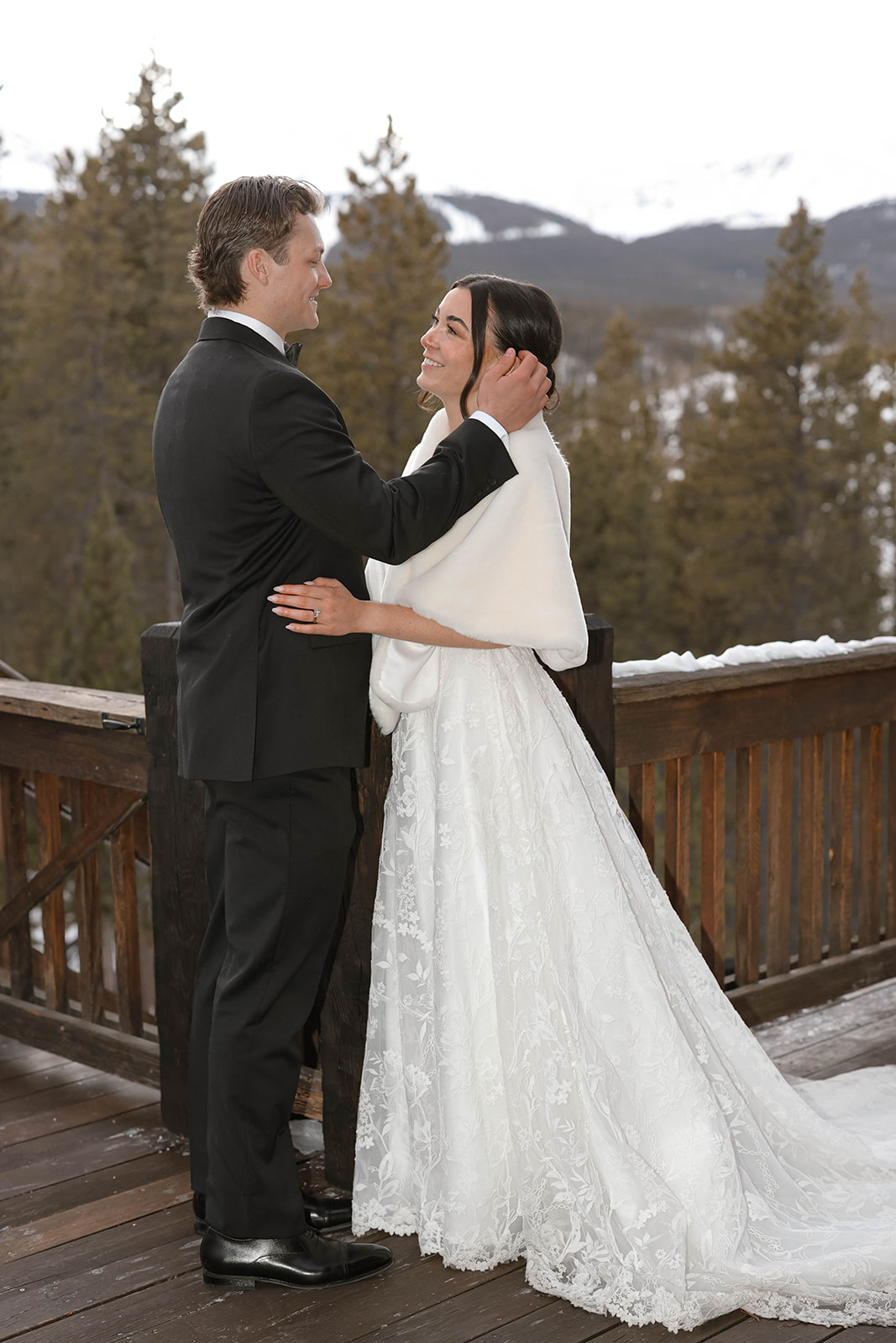 Bride and groom standing close together on a snowy wooden deck overlooking pine trees and distant mountains, the bride’s lace train and shawl flowing gently behind her.