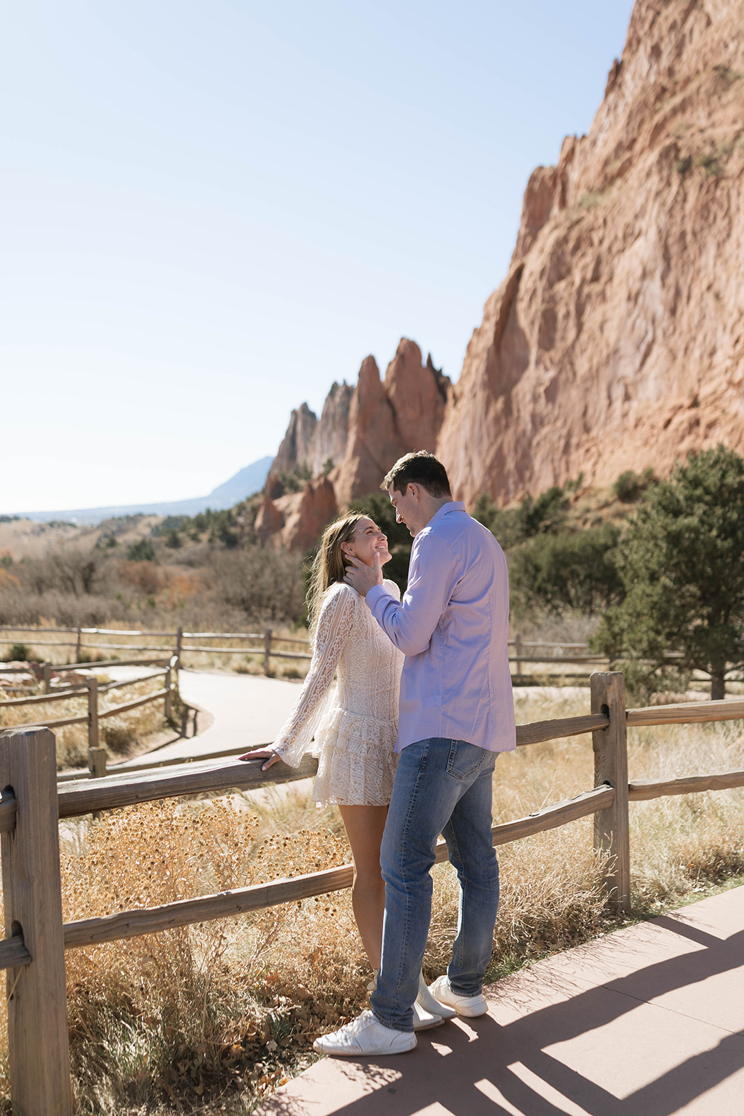 Couple standing close and gazing at each other near red rock formations and a wooden fence, the woman leaning back as he gently holds her face.