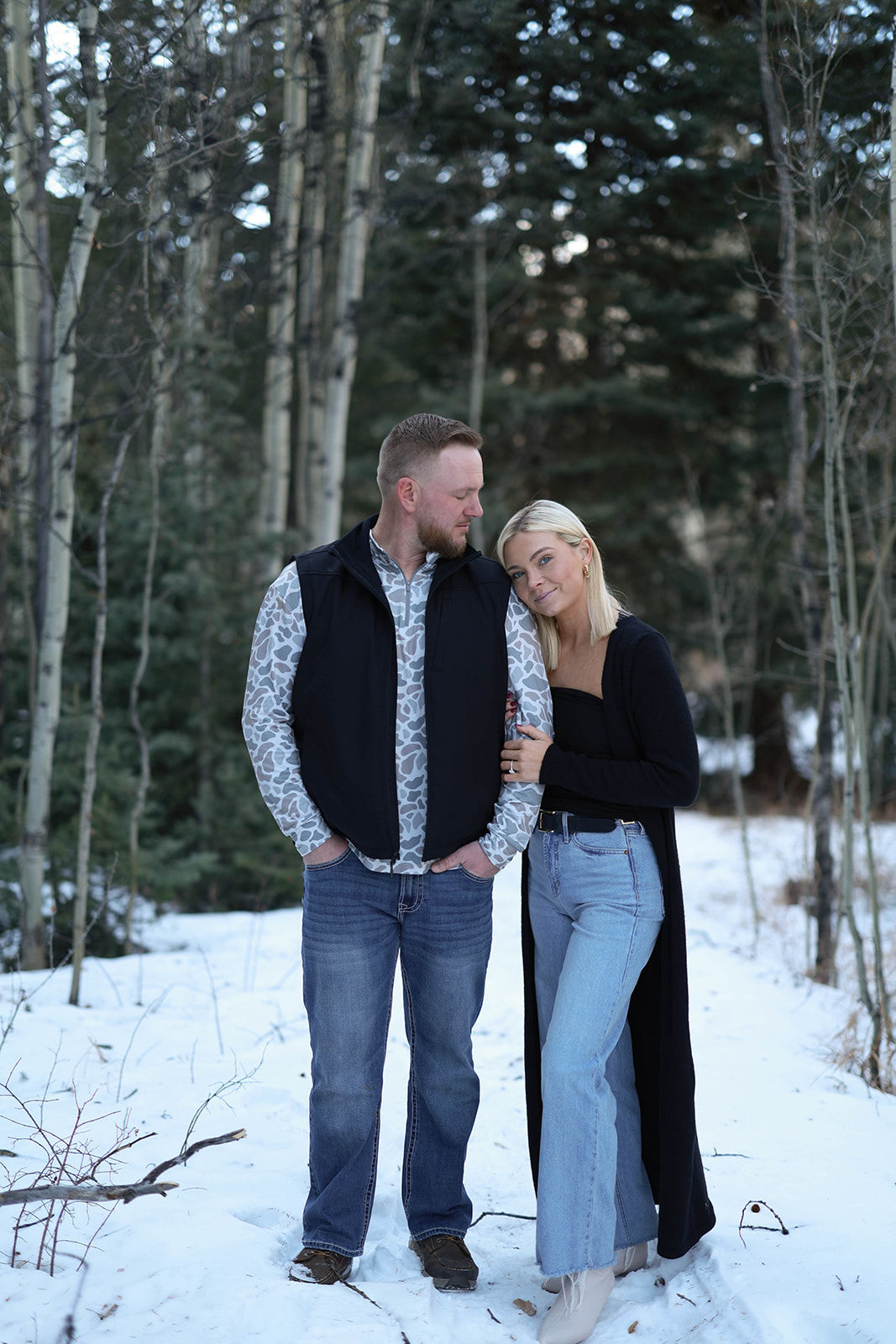 Couple standing close together in a snowy forest, the woman leaning into her partner’s shoulder as they smile softly among tall evergreen trees.