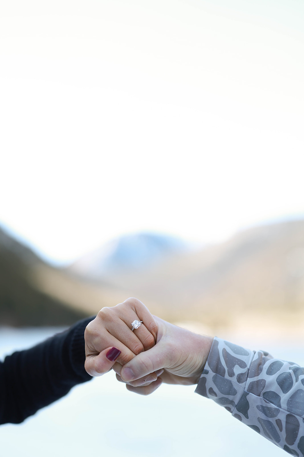 Detail shot of two hands touching knuckles, highlighting a diamond engagement ring with blurred mountains and water in the background.