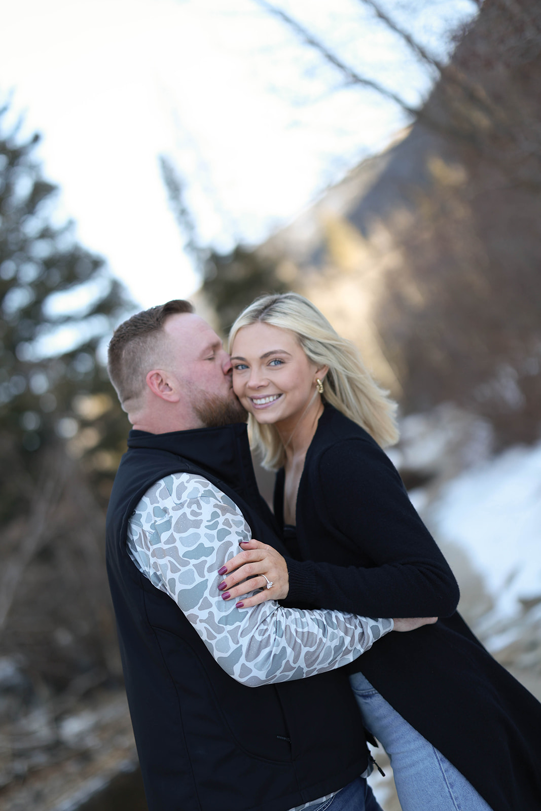 A couple embraces during a mountain proposal, with the man kissing the woman’s cheek as she smiles toward the camera, surrounded by snow, evergreen trees, and soft winter light.