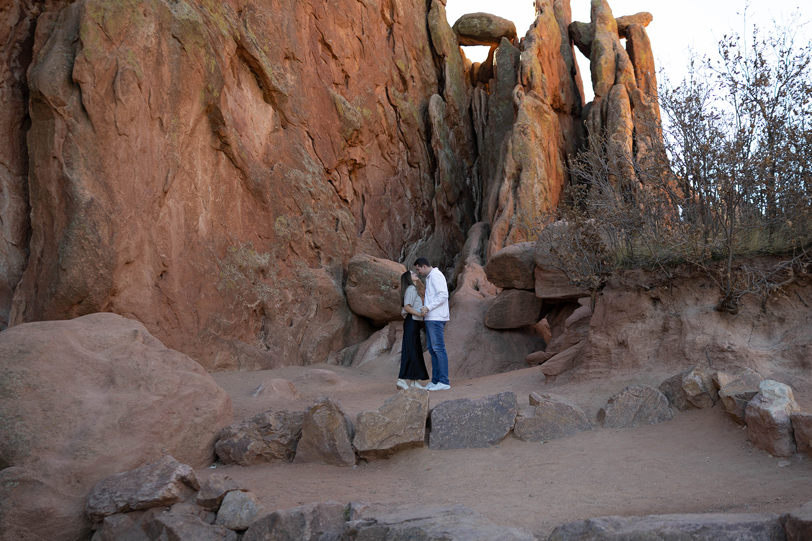 Wide shot of a couple standing together in a rocky canyon area with towering red stone walls, holding hands in a quiet, intimate moment.
