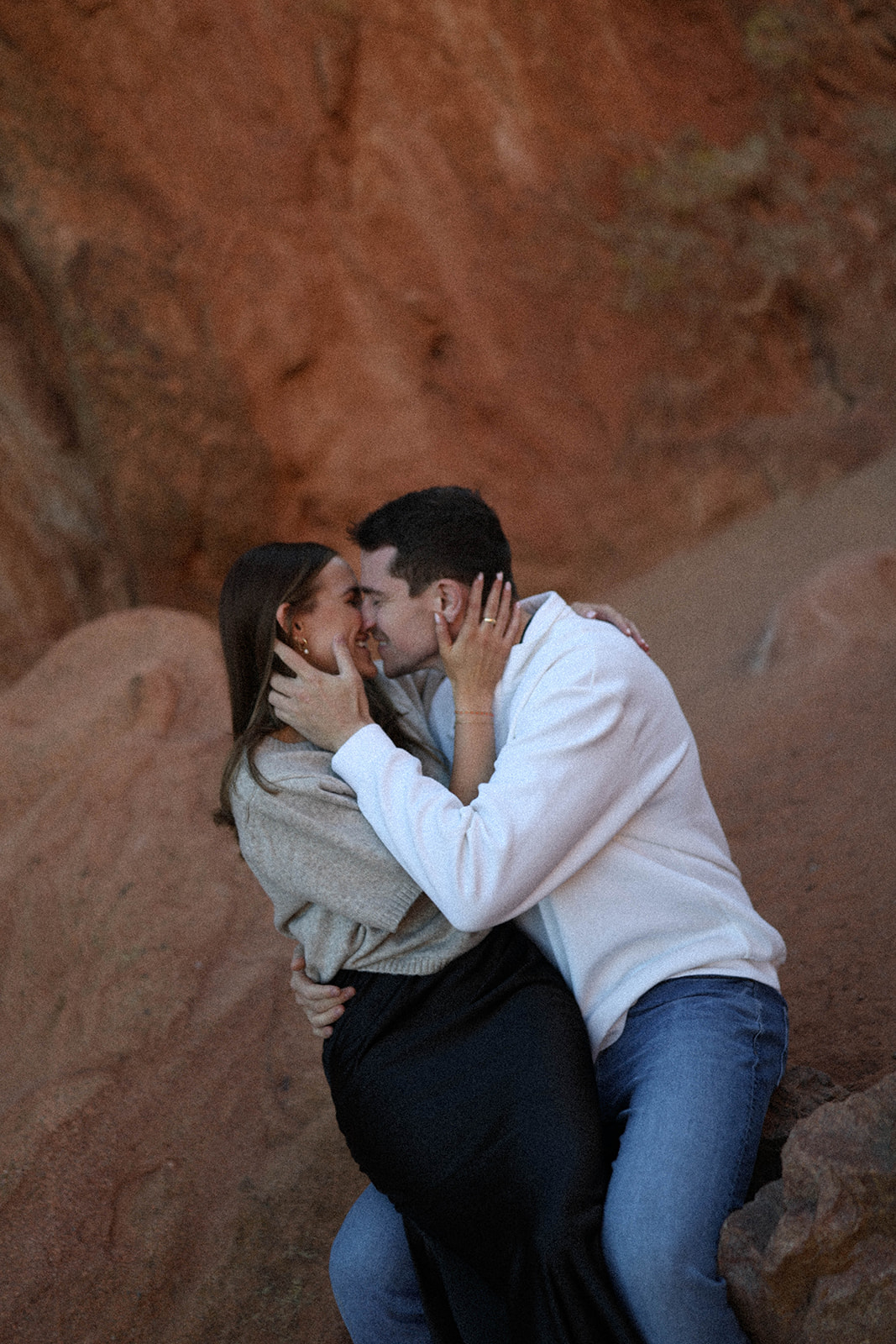 Couple kissing and holding each other against warm-toned red rock cliffs, an intimate moment captured after their mountain proposal.