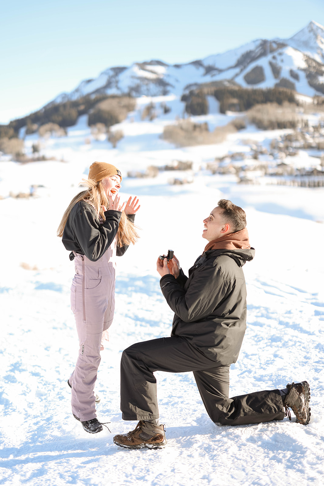 Man proposing on one knee in a snow-covered alpine landscape while his partner reacts with surprise, hands raised, surrounded by bright winter light during a mountain proposal.