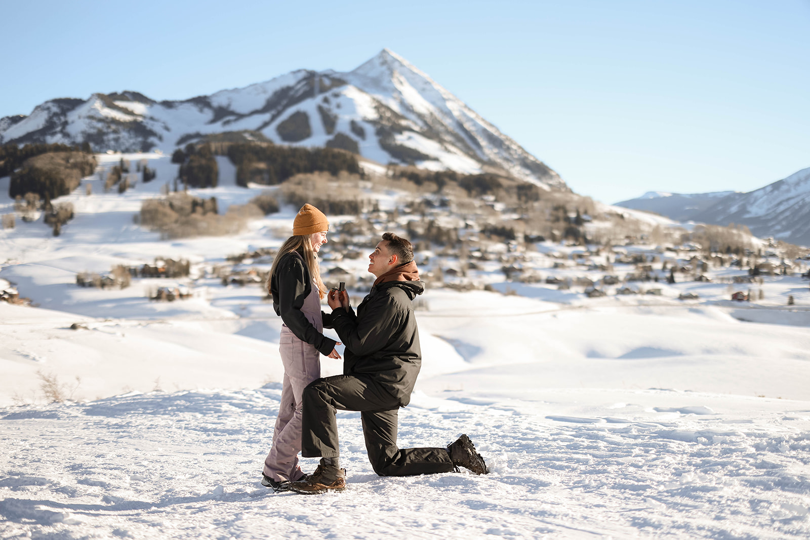 A man kneels in the snow holding a ring box while proposing to his partner during a mountain proposal, with snow-covered peaks and a small alpine town in the background.