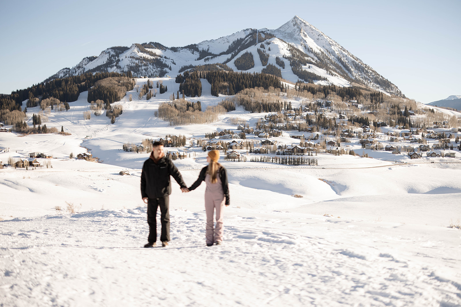 A couple walks hand in hand through a snowy landscape with a dramatic mountain backdrop, documenting a joyful mountain proposal adventure.