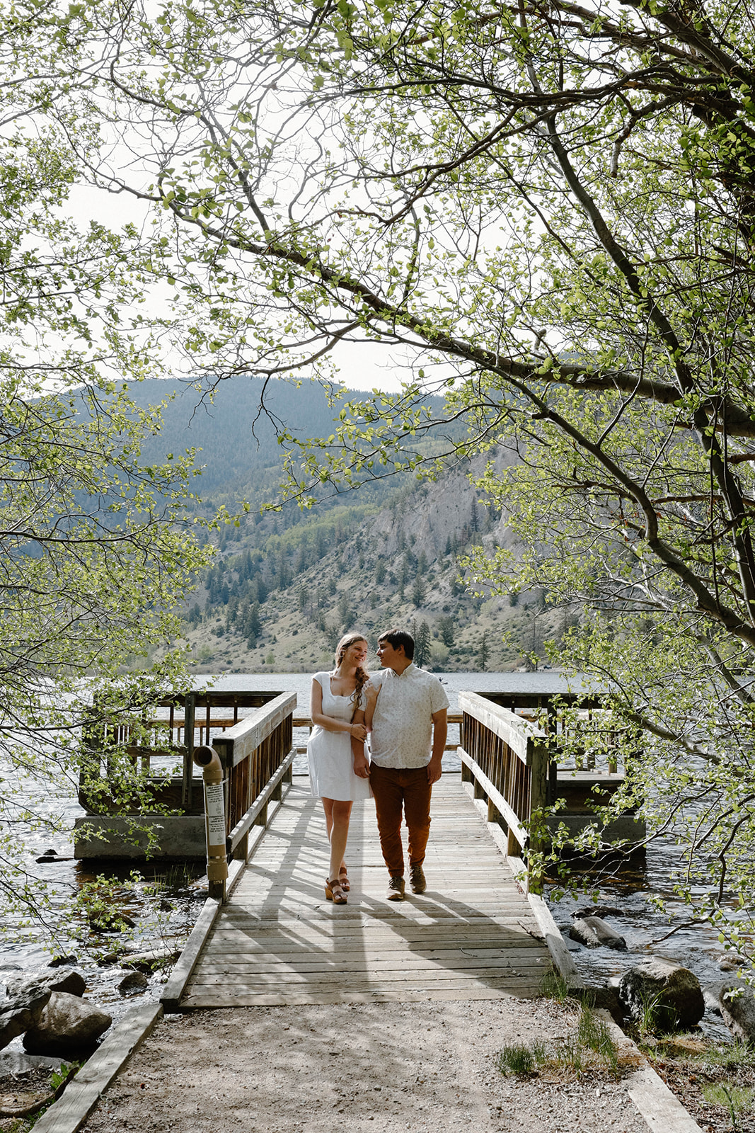 Couple walking hand in hand across a wooden dock surrounded by trees and a calm lake, with mountains rising in the background during a peaceful mountain proposal setting.