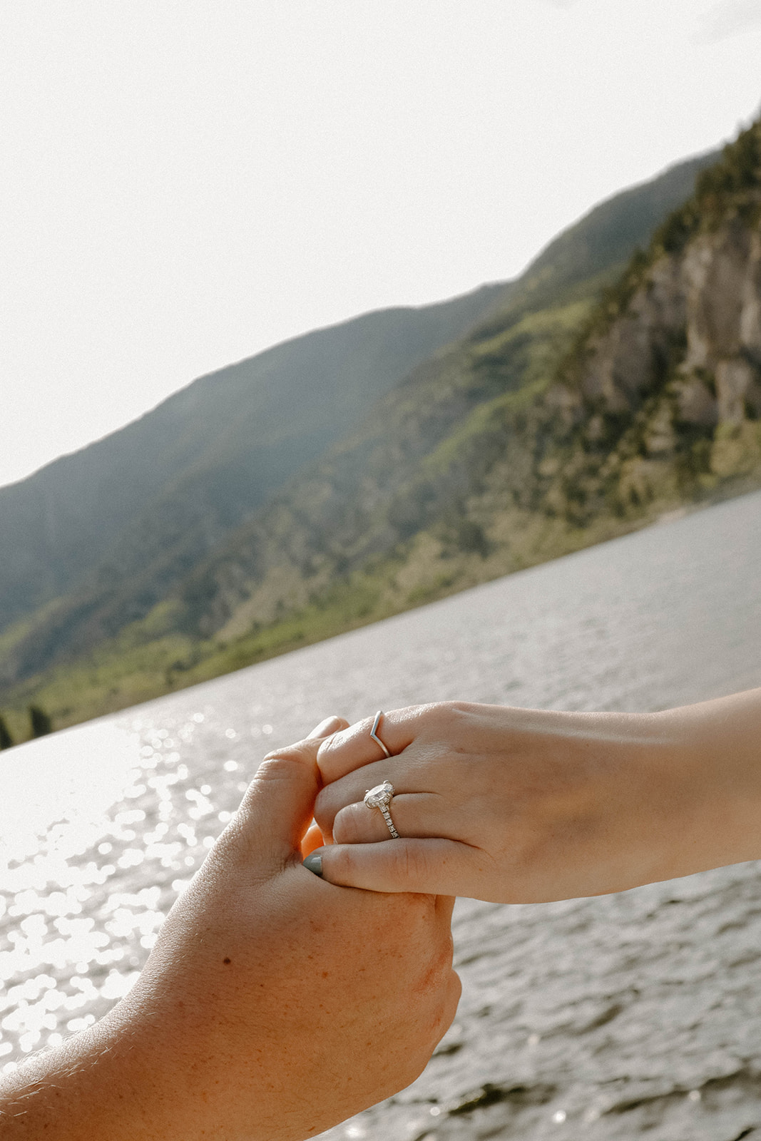 Close-up of hands holding together near a mountain lake, highlighting an engagement ring sparkling in the sunlight.