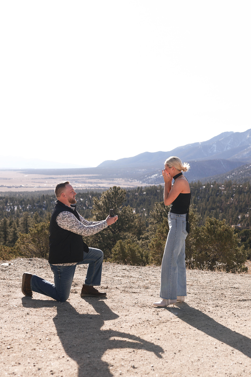 Man kneeling on one knee holding an open ring box while proposing to his partner at a scenic overlook with pine trees and mountain views behind them, a heartfelt mountain proposal moment.