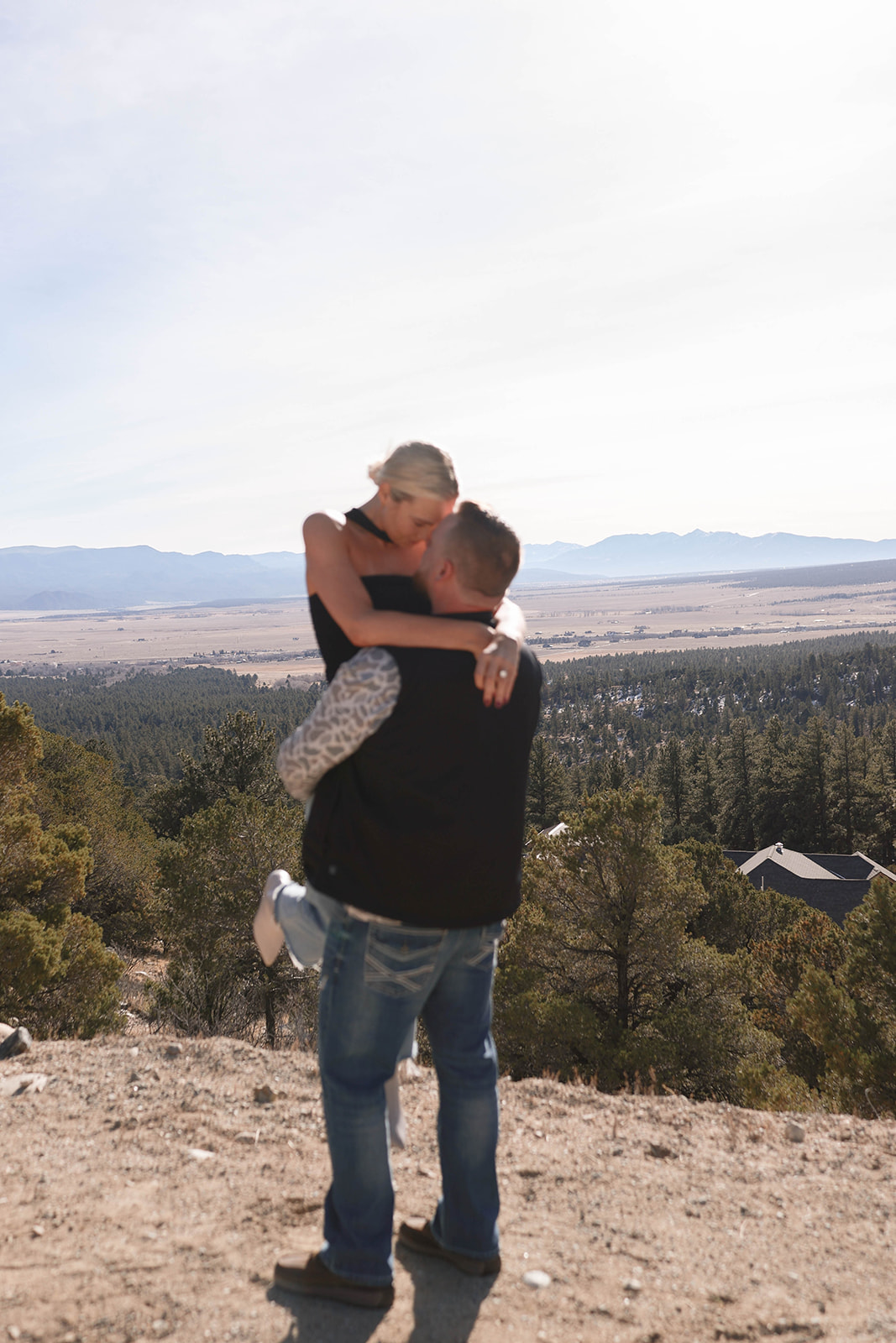 A couple shares an intimate moment on a scenic overlook as the man lifts the woman in his arms, with expansive forested hills and distant mountains stretching across the horizon behind them.