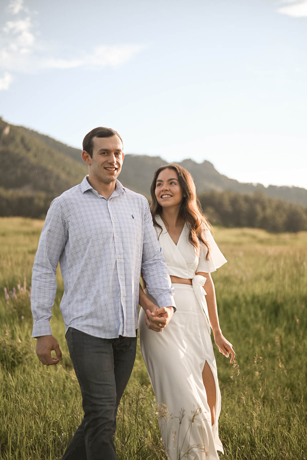 A couple strolls through an open grassy field with mountains in the background, wearing a soft white dress and light button-up shirt for a romantic, effortless look.