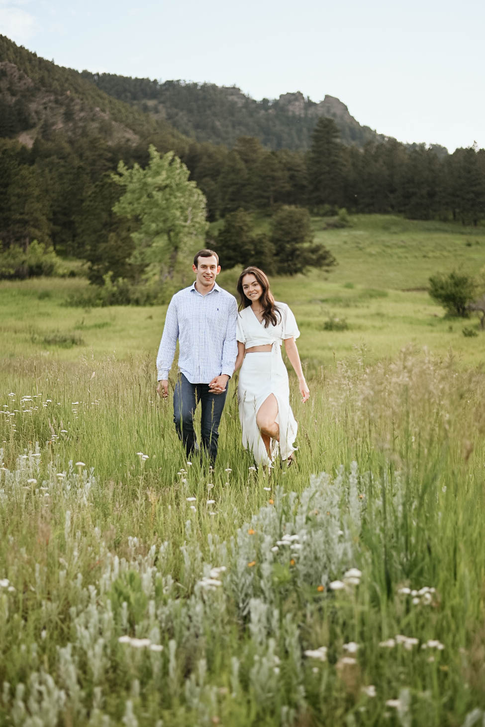 A couple walking hand in hand through tall grass in a mountain field, celebrating their engagement after a mountain proposal.