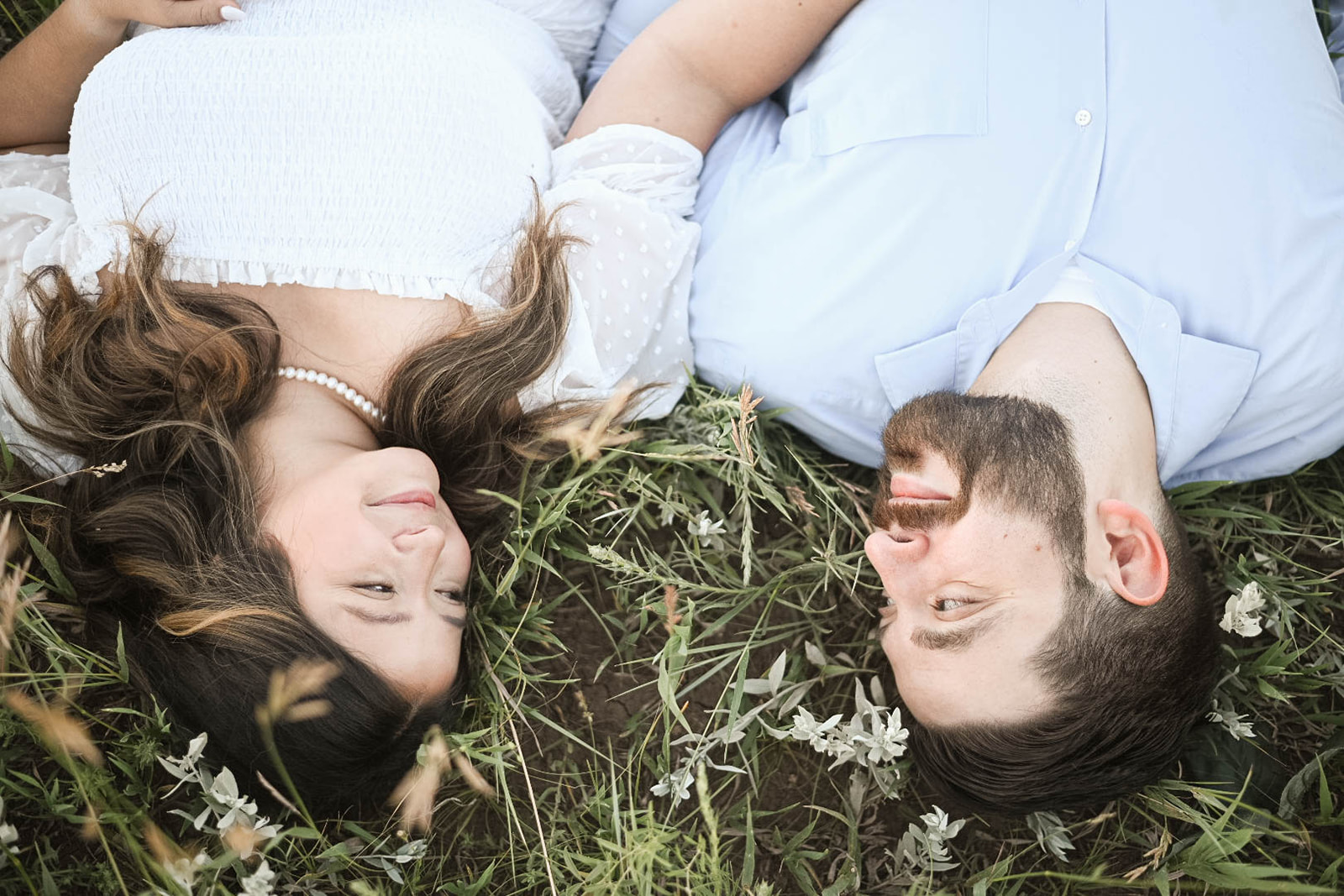 A couple lies on their backs in a grassy field, facing each other and smiling softly, capturing a quiet engagement portrait moment.