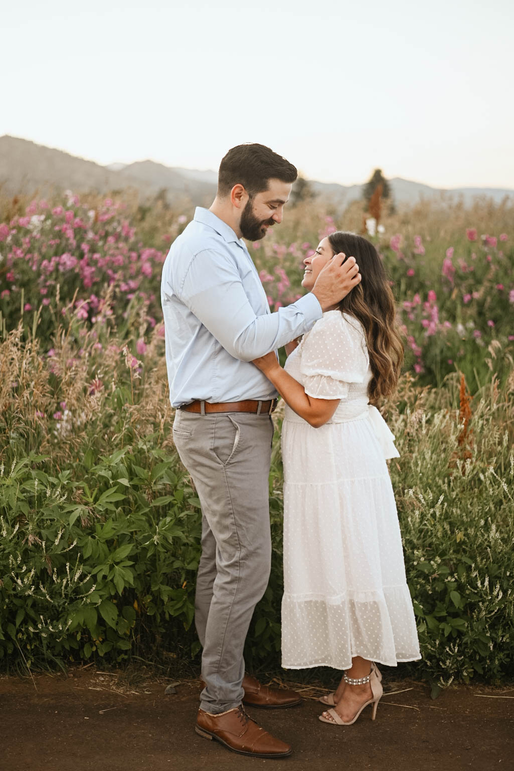 A couple standing close together in a wildflower meadow at sunset, smiling at each other during a quiet mountain proposal moment.