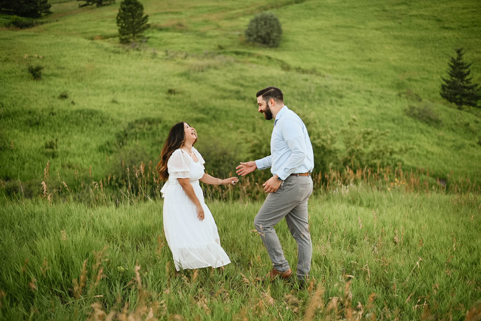 Couple laughing and reaching for each other in a grassy field during a joyful outdoor mountain proposal moment.