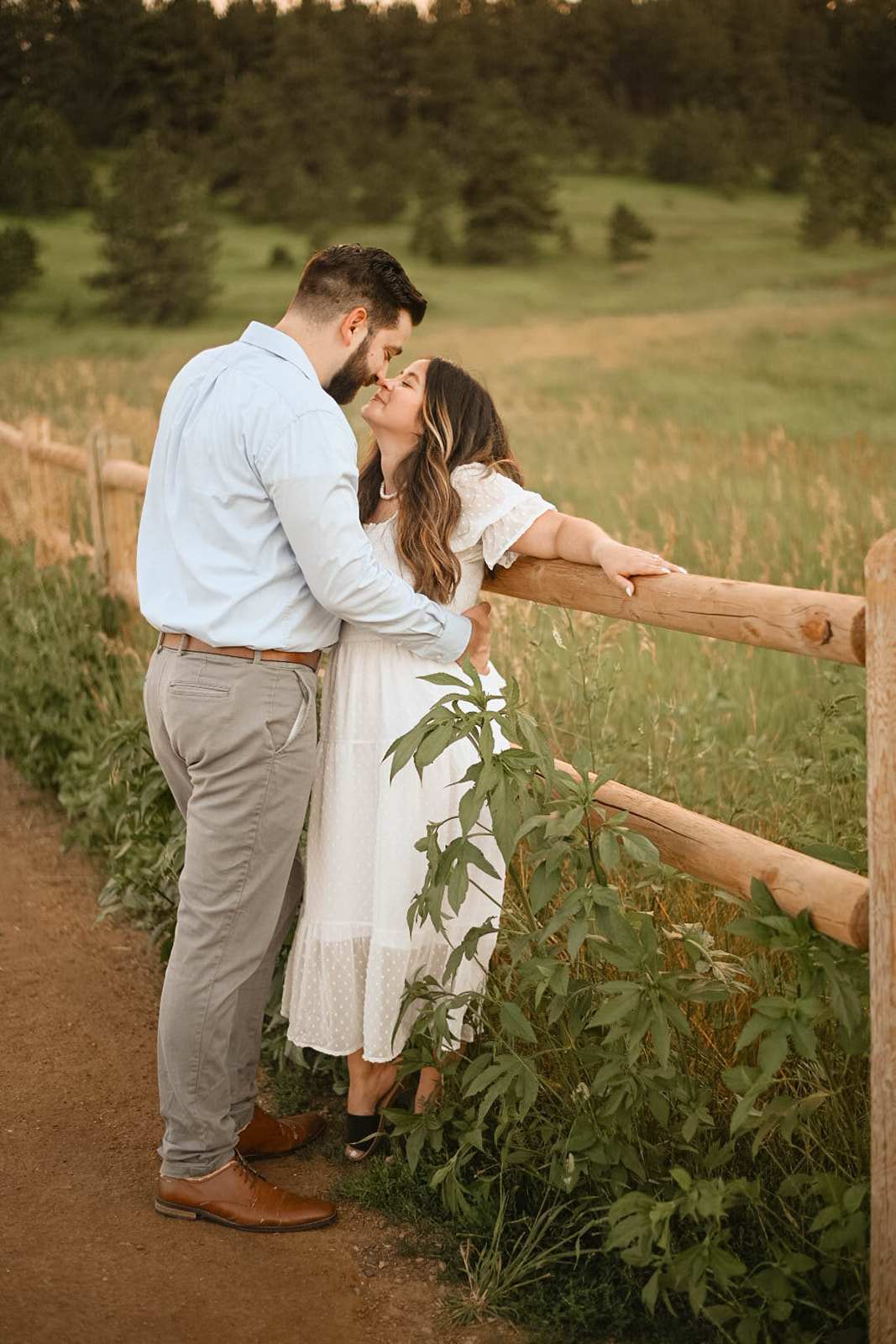 Couple standing by a wooden fence in an open field, sharing a quiet kiss during a romantic mountain proposal at golden hour.