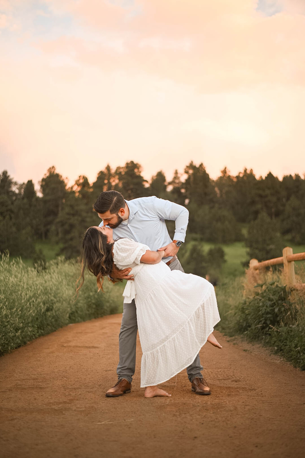 Groom dipping his partner into a kiss on a dirt path at sunset, capturing post-proposal joy in a natural outdoor setting.