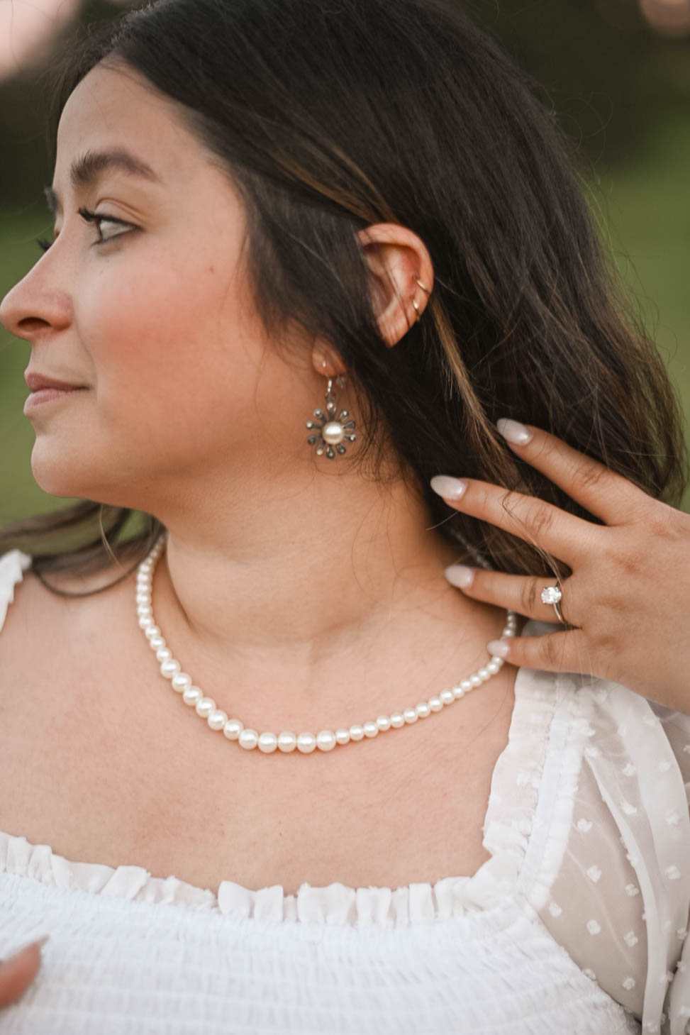 Close-up side profile of a woman wearing pearl earrings and a necklace, with her engagement ring visible as she tucks her hair behind her ear.