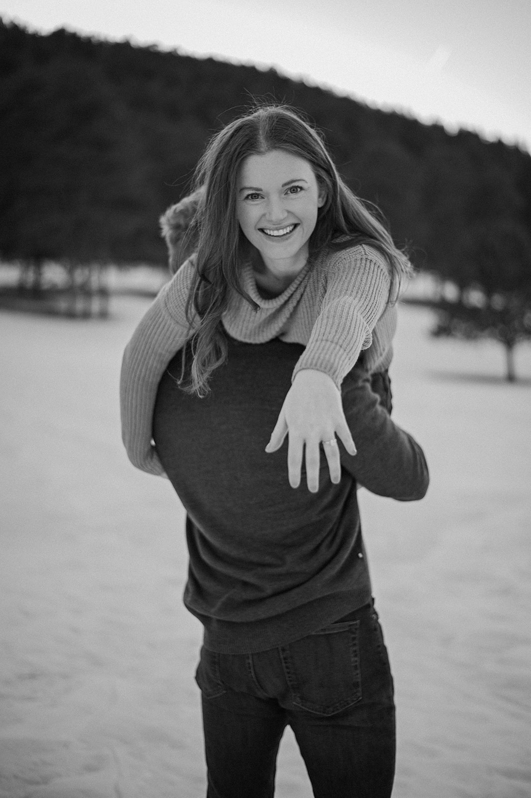 Black-and-white photo of a woman smiling while being carried piggyback in a snowy field, showing her engagement ring extended toward the camera.