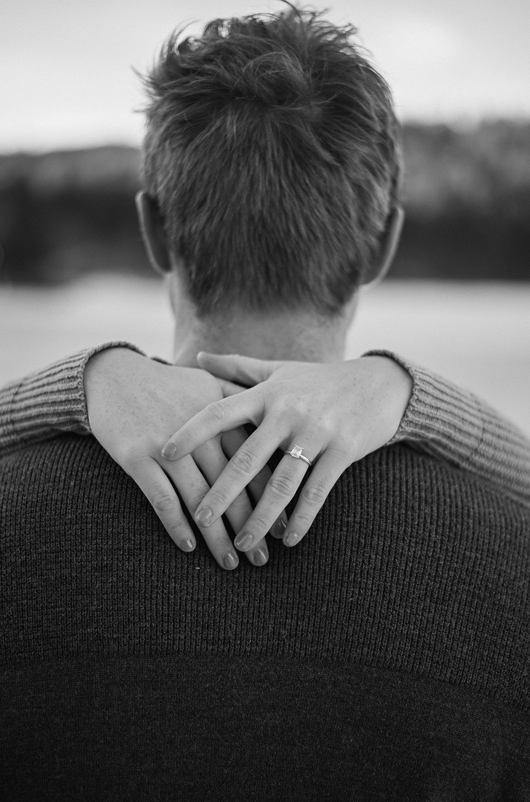 Black-and-white close-up from behind of a woman’s hands resting on her partner’s shoulders, highlighting her engagement ring after a mountain proposal.