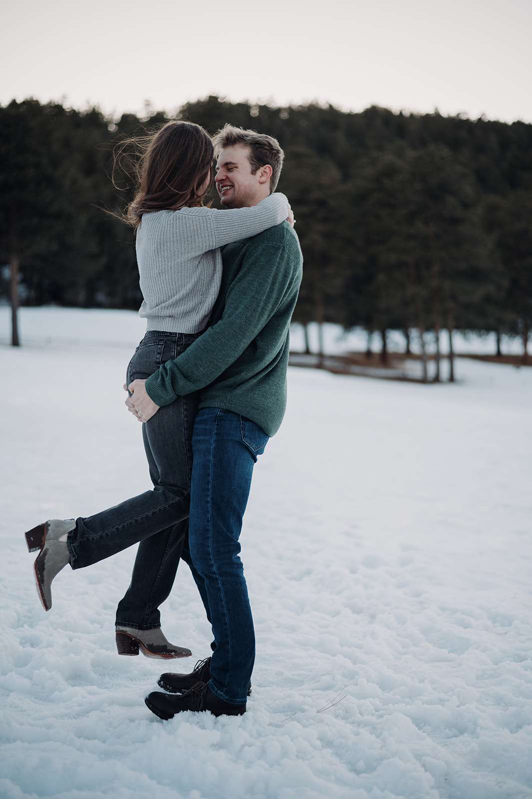 A couple embraces in the snow as one partner lifts the other, sharing a joyful moment during a winter mountain proposal session surrounded by pine trees.