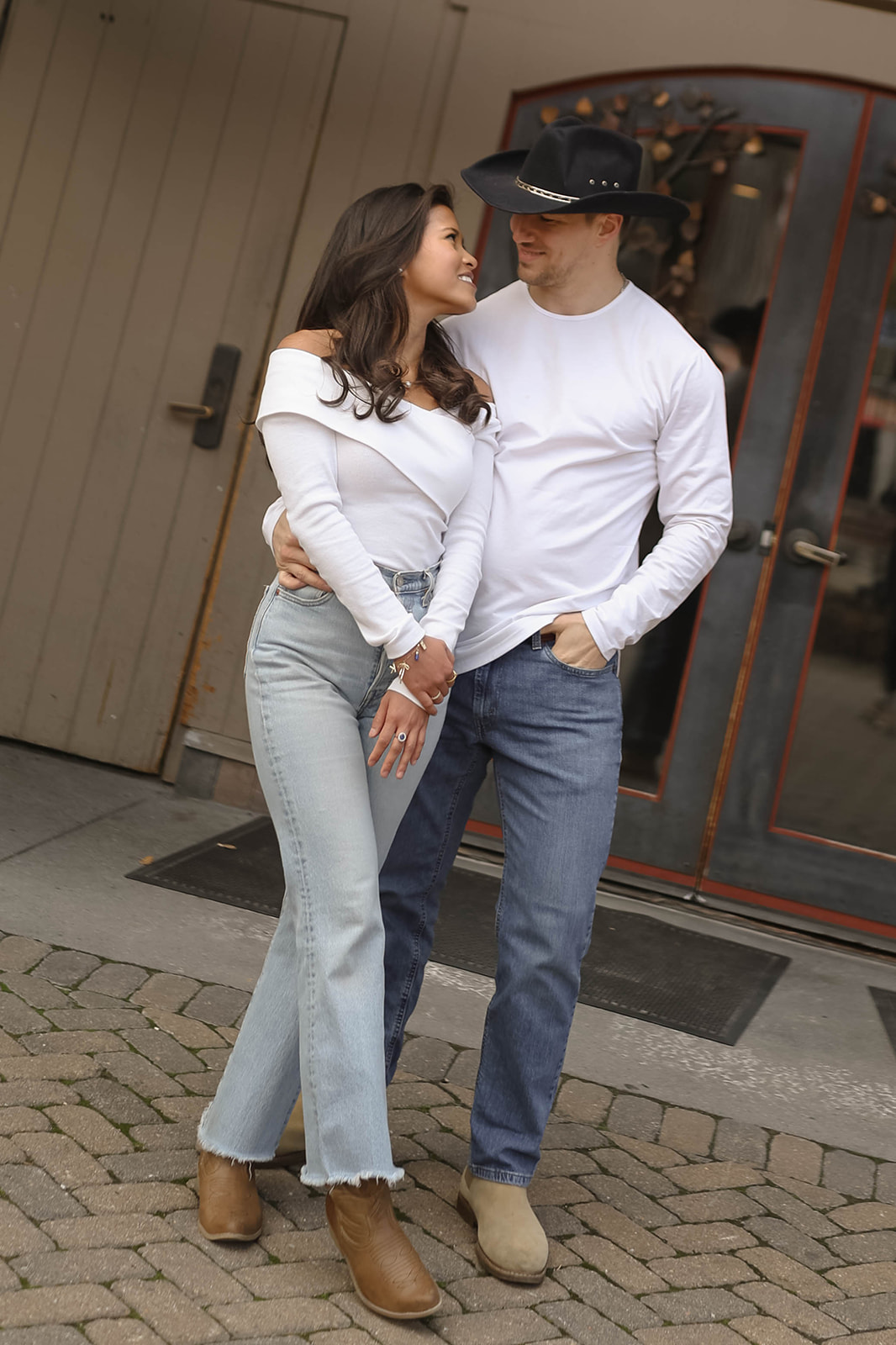 Couple standing outside a doorway in black cowboy hats, with her in an off-the-shoulder white top and light jeans and him in a white long-sleeve shirt and dark jeans.