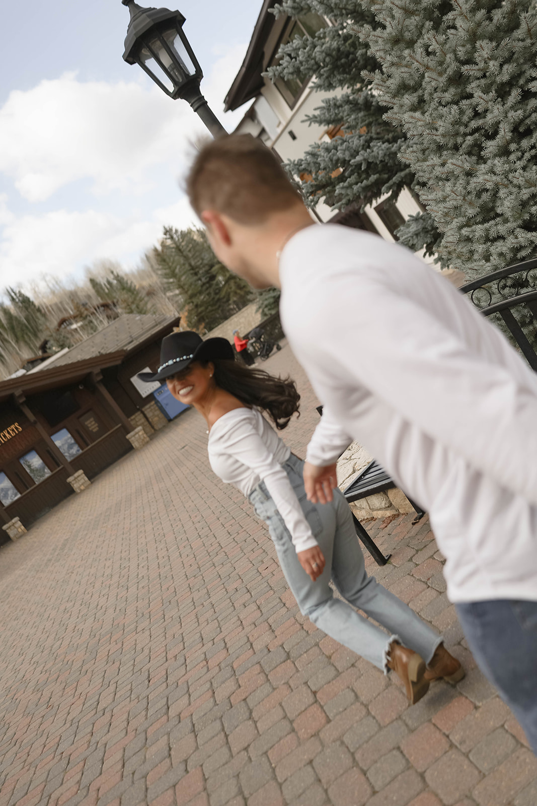 A candid motion shot of a couple walking through a mountain village plaza, the woman wearing a wide-brim black hat and off-the-shoulder top, showcasing modern couples outfit ideas with western-inspired details.