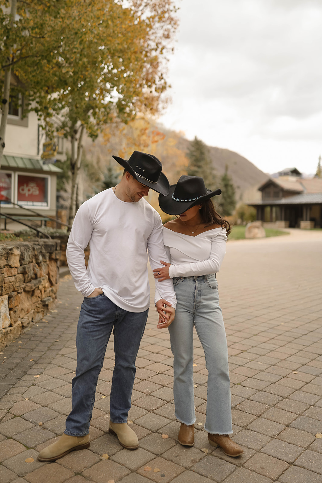 Western-style couple walking hand-in-hand on a small-town street in matching black cowboy hats, white tops, jeans, and tan boots—easy couples outfit ideas for a laid-back session.