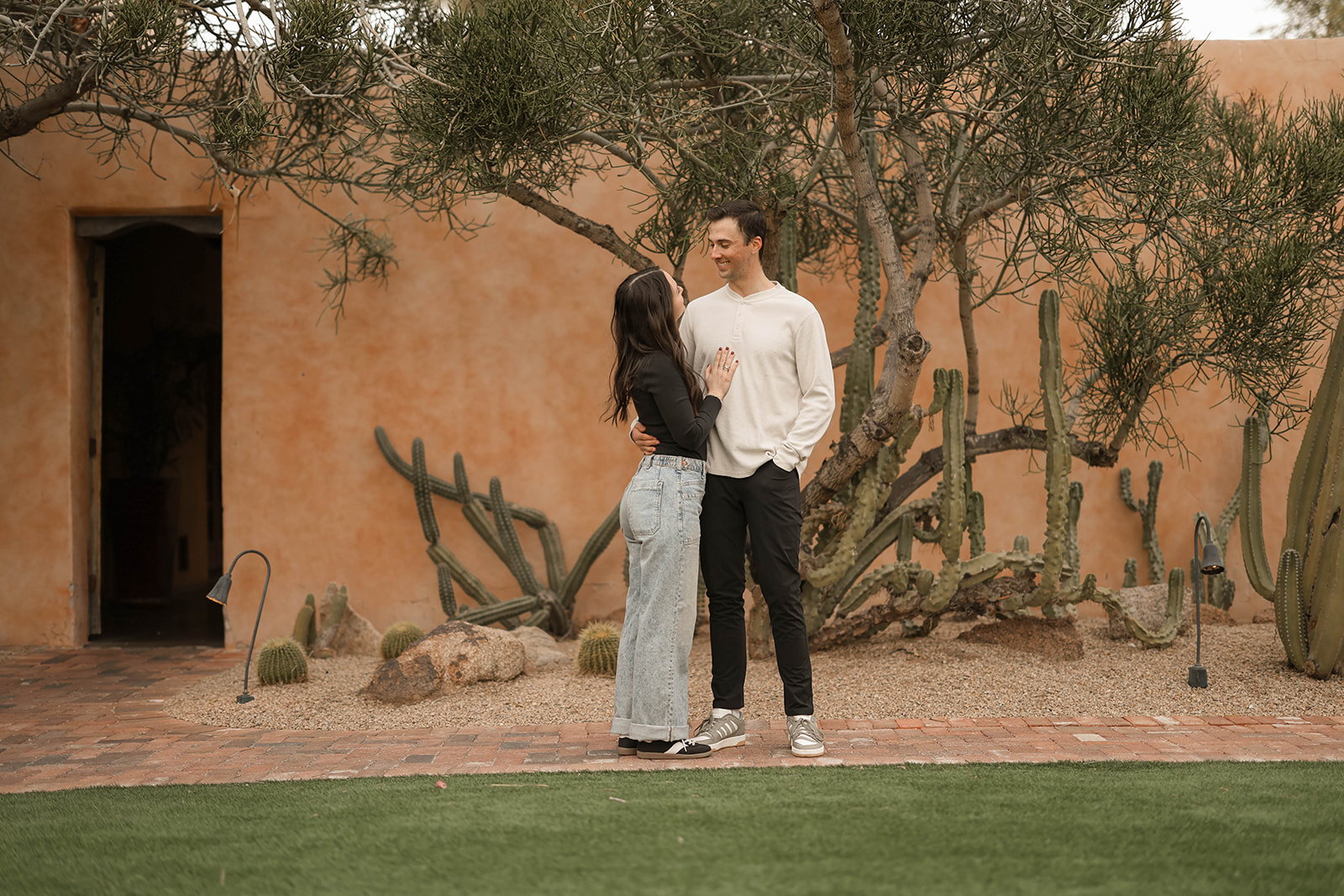 A couple stands close together in a desert courtyard with cacti and an adobe building behind them, smiling at each other while dressed in neutral, casual outfits.