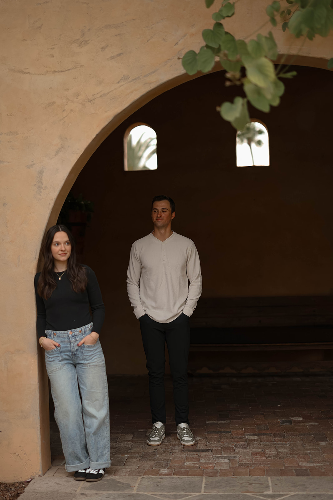 A couple stands apart beneath a large adobe archway, with the woman leaning against the wall and the man standing a few feet away, both wearing neutral tops and jeans in a quiet courtyard setting.