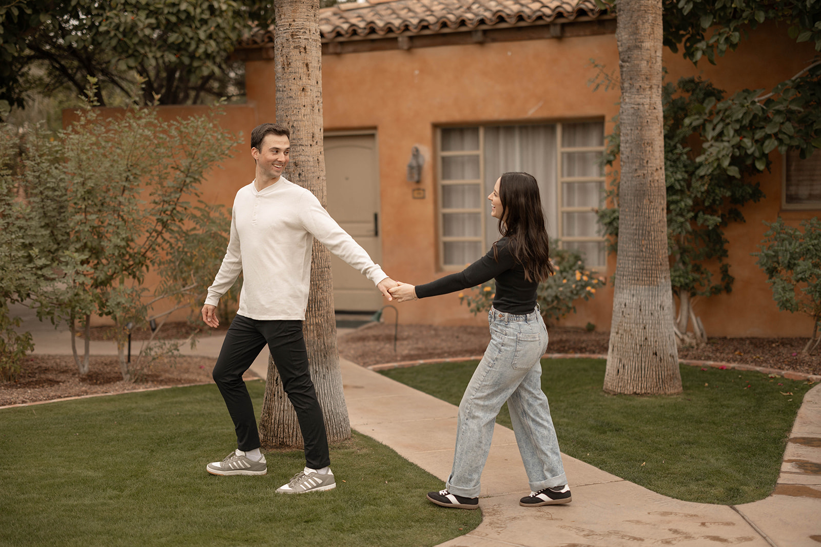 A couple walks hand in hand across a grassy courtyard framed by palm trees and a warm-toned stucco building, dressed in neutral tops and casual denim for timeless couples photos.