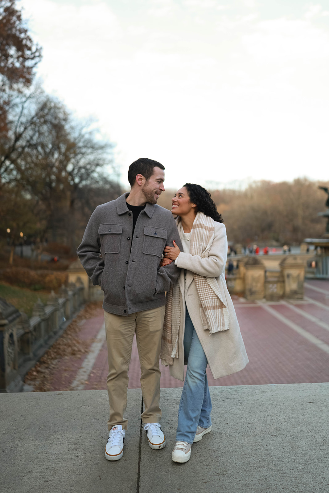 Couple walking arm-in-arm across a bridge in Central Park, wearing neutral-toned coats, scarves, and casual sneakers — cozy fall couples outfit ideas for a relaxed city engagement session.