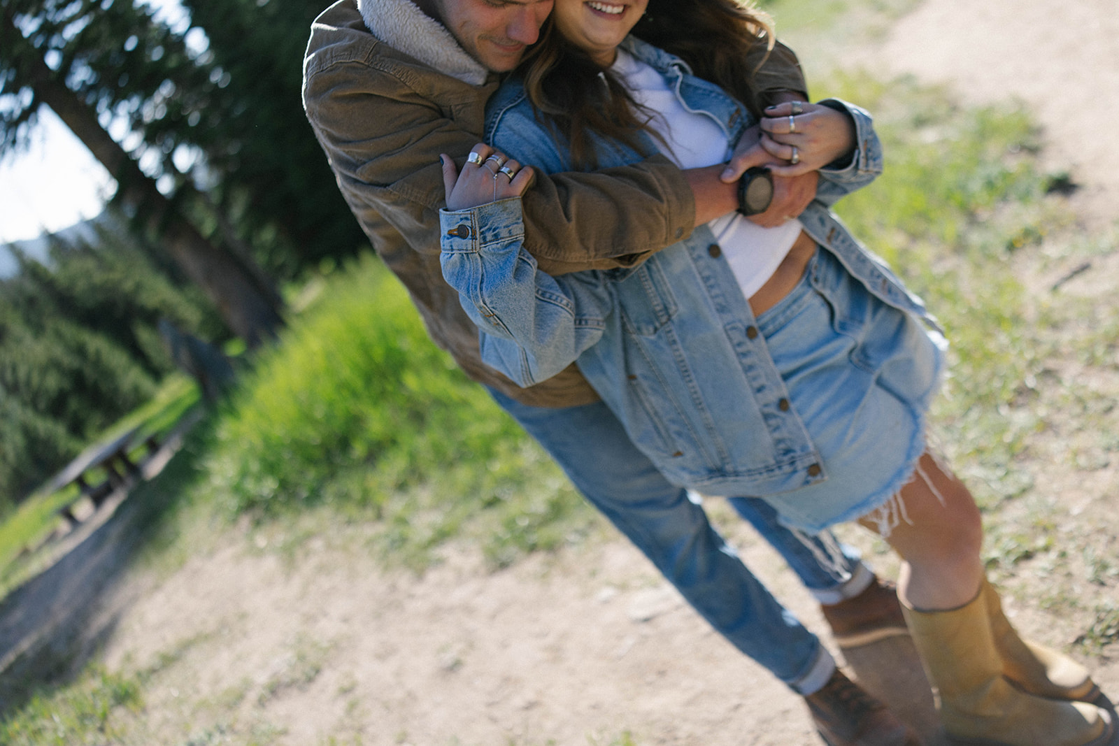 A couple embraces outdoors as the man wraps his arms around the woman from behind, both wearing denim layers while standing on a grassy path in soft sunlight.