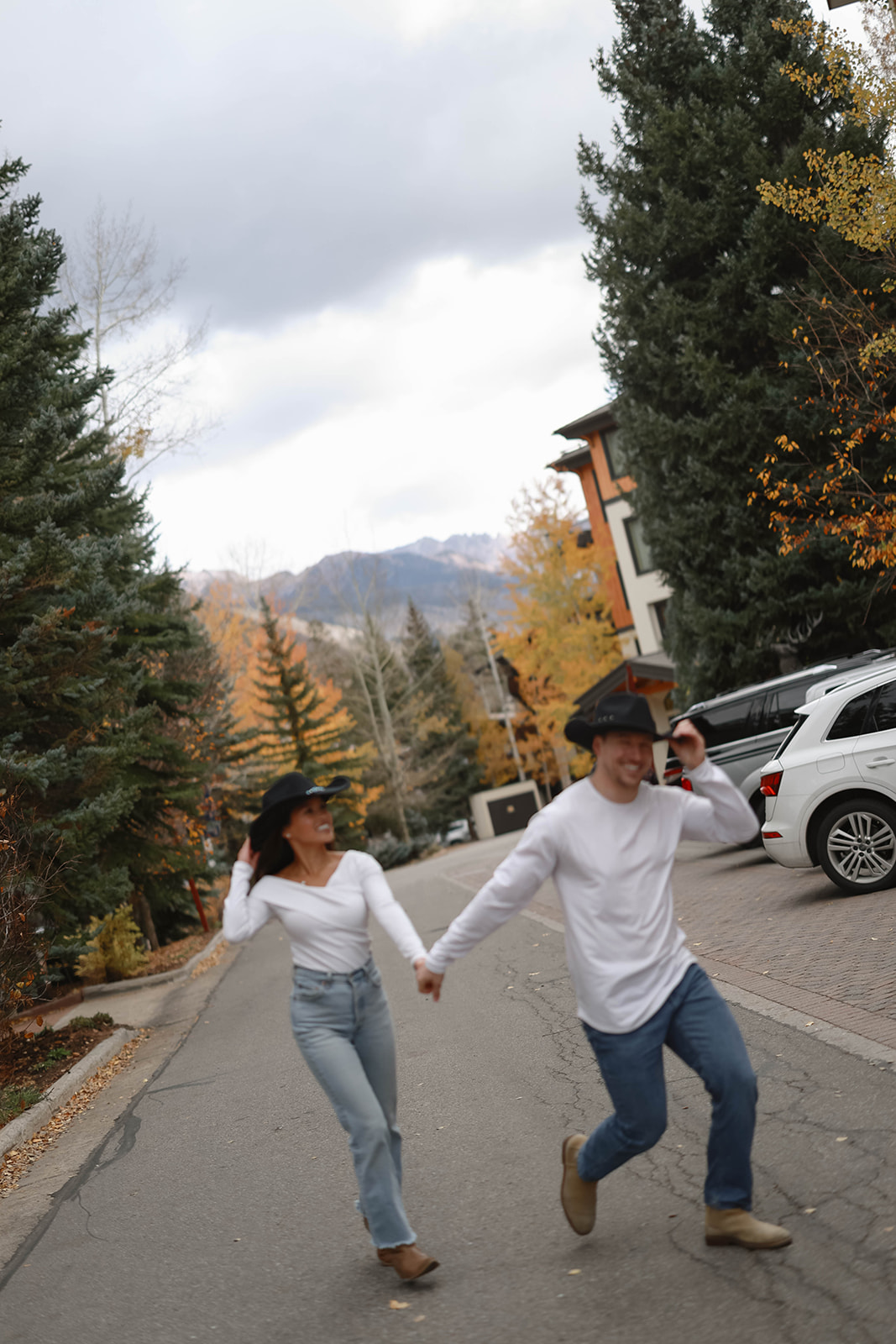 A couple runs hand in hand down a paved mountain street lined with evergreen trees and autumn leaves, both wearing white tops, jeans, and black hats.
