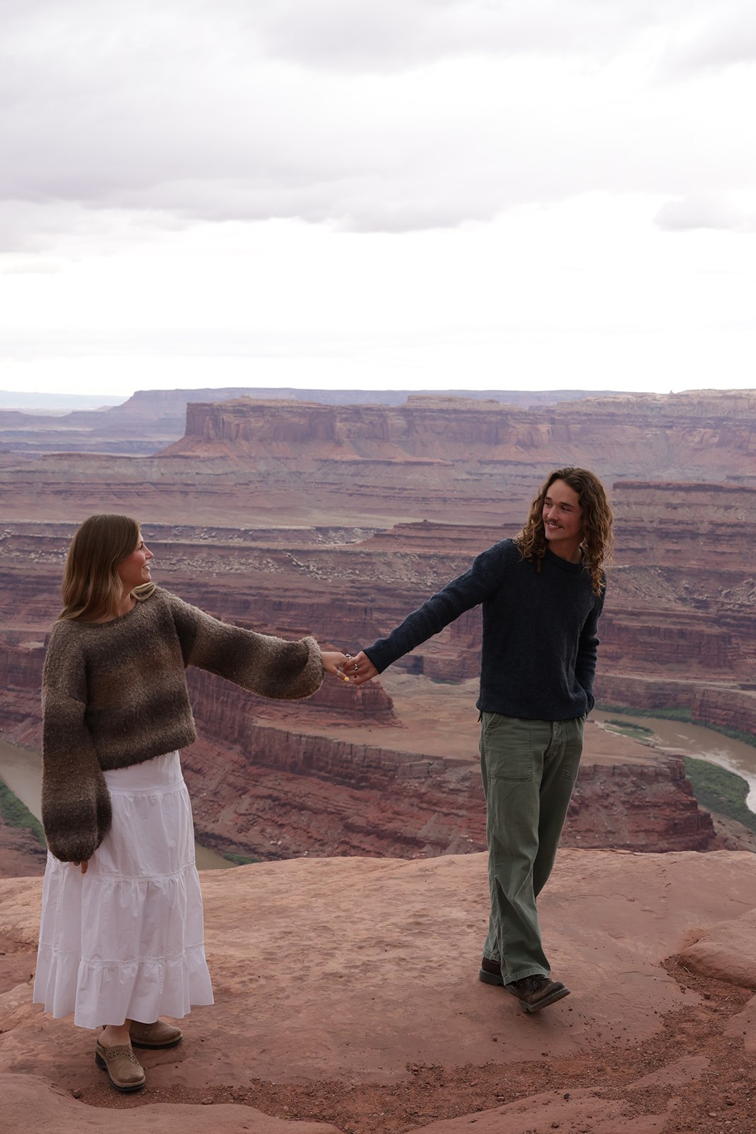 A couple holds hands at a canyon overlook, smiling at each other with layered red rock cliffs and a winding river visible in the distance.