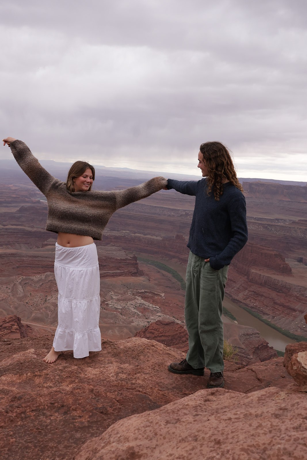 A couple stands at the edge of a canyon overlook holding hands, with the woman lifting her arms wide and the layered red rock canyon stretching behind them under an overcast sky.