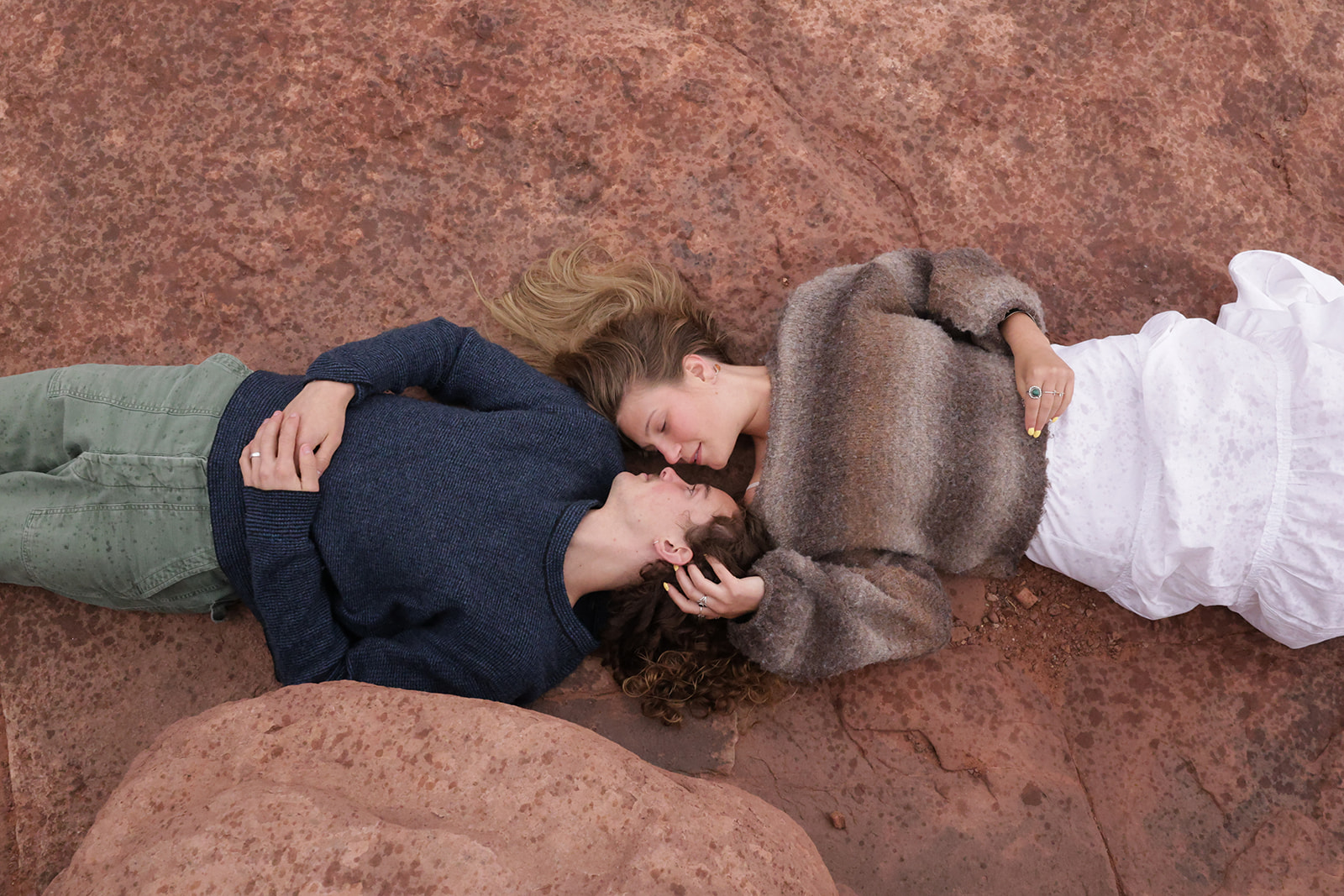 Overhead image of a couple lying on red rock terrain, wrapped in cozy layers and soft fabrics, creating a warm and intimate engagement portrait.