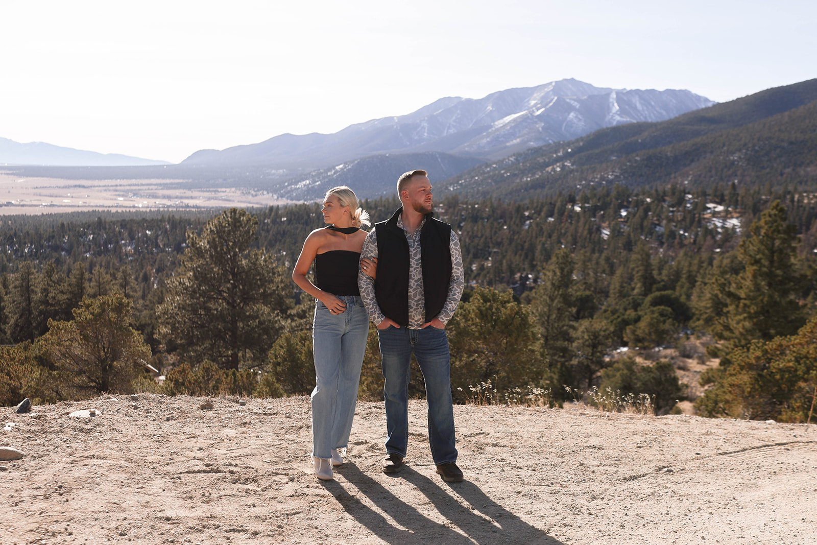 Couple standing on a mountain overlook with a wide forest-and-peak view, wearing jeans with a black strapless top and a black vest layered over a patterned shirt.