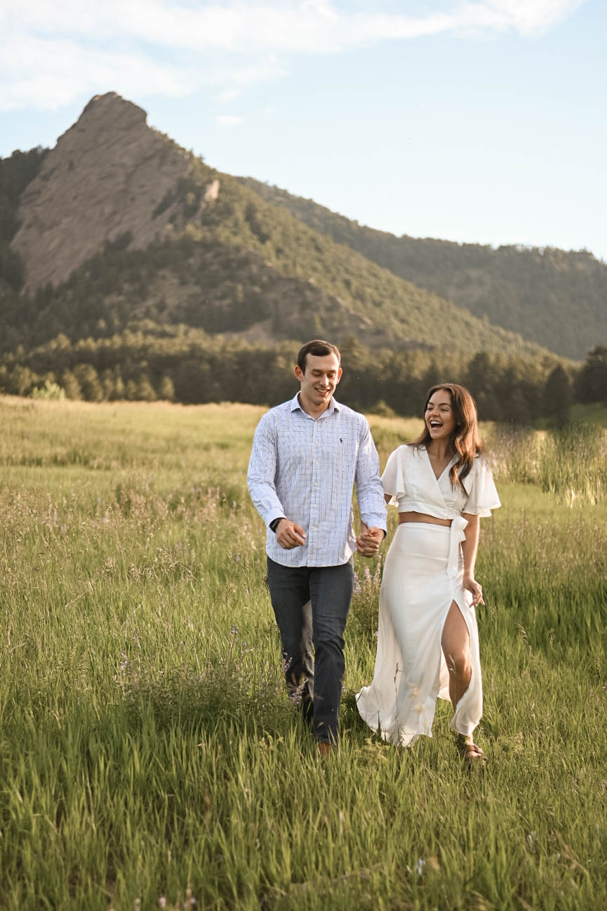 Couple laughing while walking through a green meadow with a mountain backdrop, wearing a light button-down shirt and a white two-piece set.
