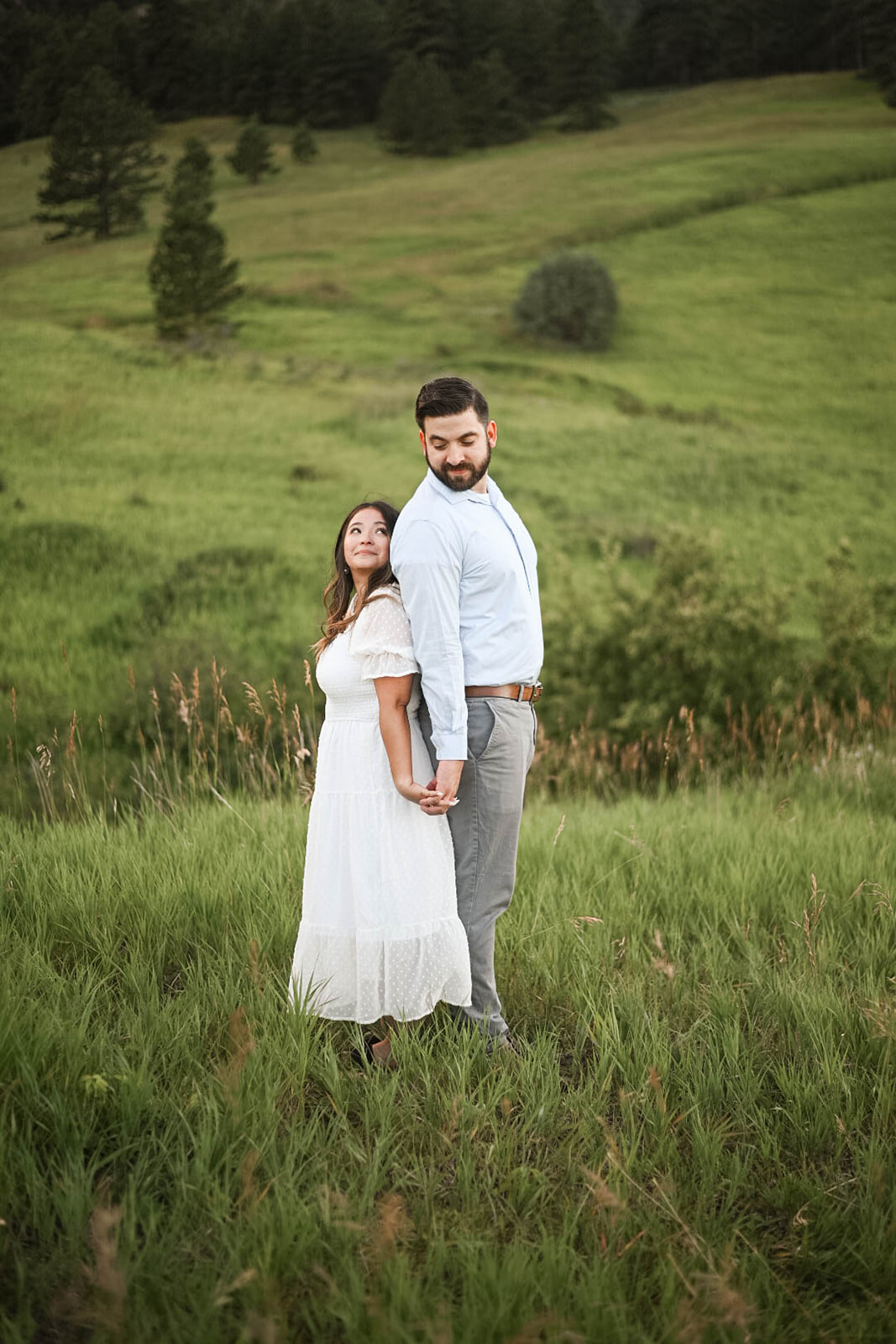 Couple standing back-to-back holding hands in a grassy field, with her in a white dress and him in a light blue shirt and gray pants—classic couples outfit ideas for outdoor photos.