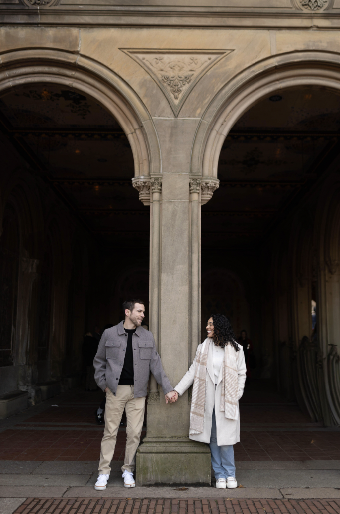 Couple holding hands under a stone archway in Central Park, framed by symmetrical architecture and soft natural light, creating a timeless and romantic engagement photo.