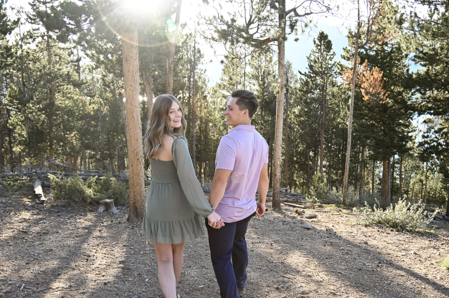 Couple walking hand-in-hand through a sunlit pine forest trail, wearing soft neutral tones and casual layers that showcase effortless couples outfit ideas for an outdoor engagement session.