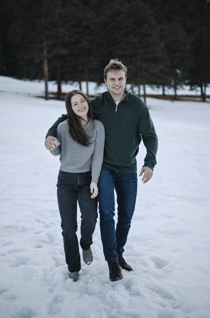 Couple walking together through fresh snow in a winter forest, dressed in cozy sweaters, denim, and boots for a playful and romantic cold-weather engagement photo.