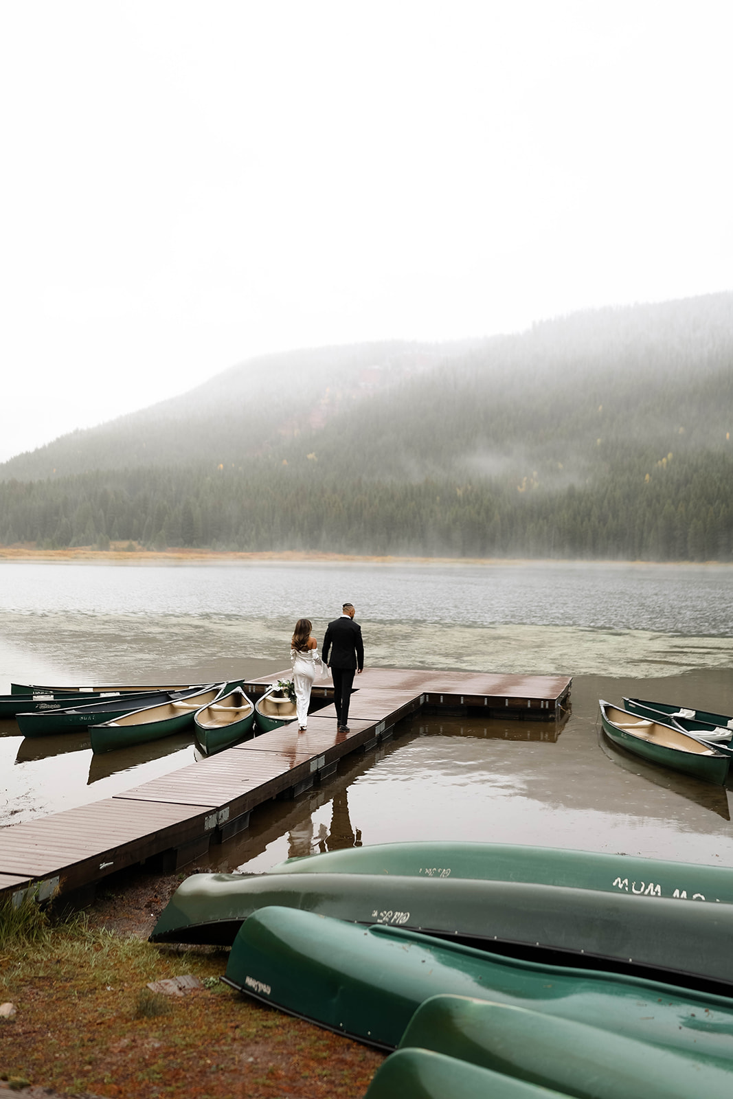 Bride and groom walk down a wooden dock toward a misty lake, surrounded by canoes and pine-covered mountains in soft autumn rain.