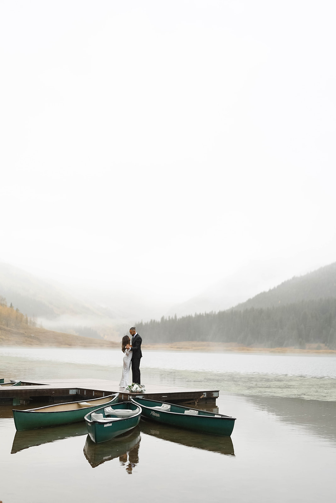 Wide shot of a couple standing together on a dock surrounded by calm lake water, green canoes, and misty mountain scenery during a vail elopement.