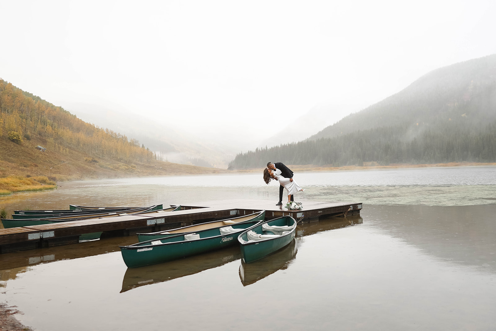 Bride and groom sharing a dipped kiss on a wooden dock beside green canoes, with fog rolling over a mountain lake in Vail, Colorado.
