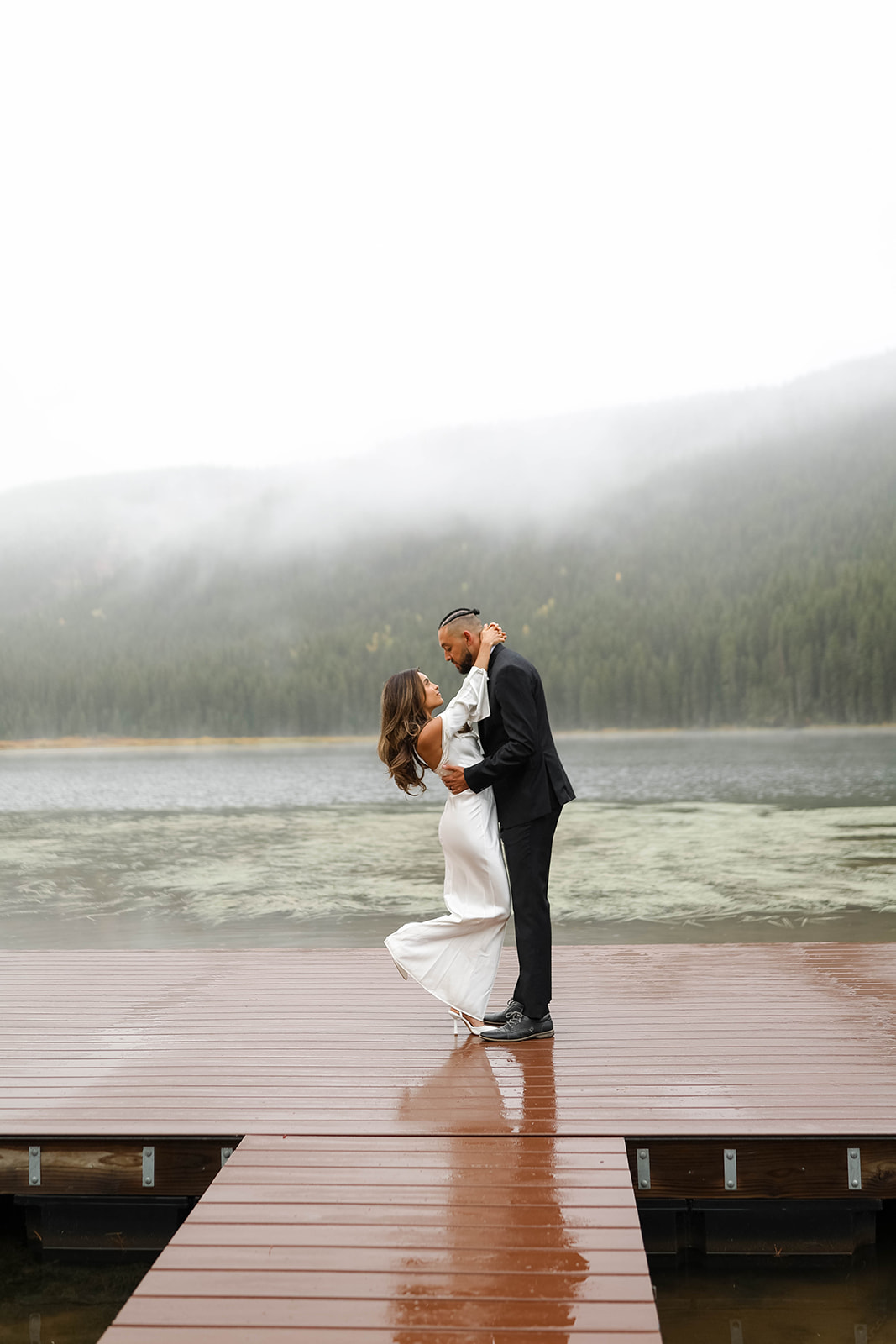 Bride and groom share an intimate kiss on a rain-soaked dock during a vail elopement, with fog drifting over the mountain lake behind them.