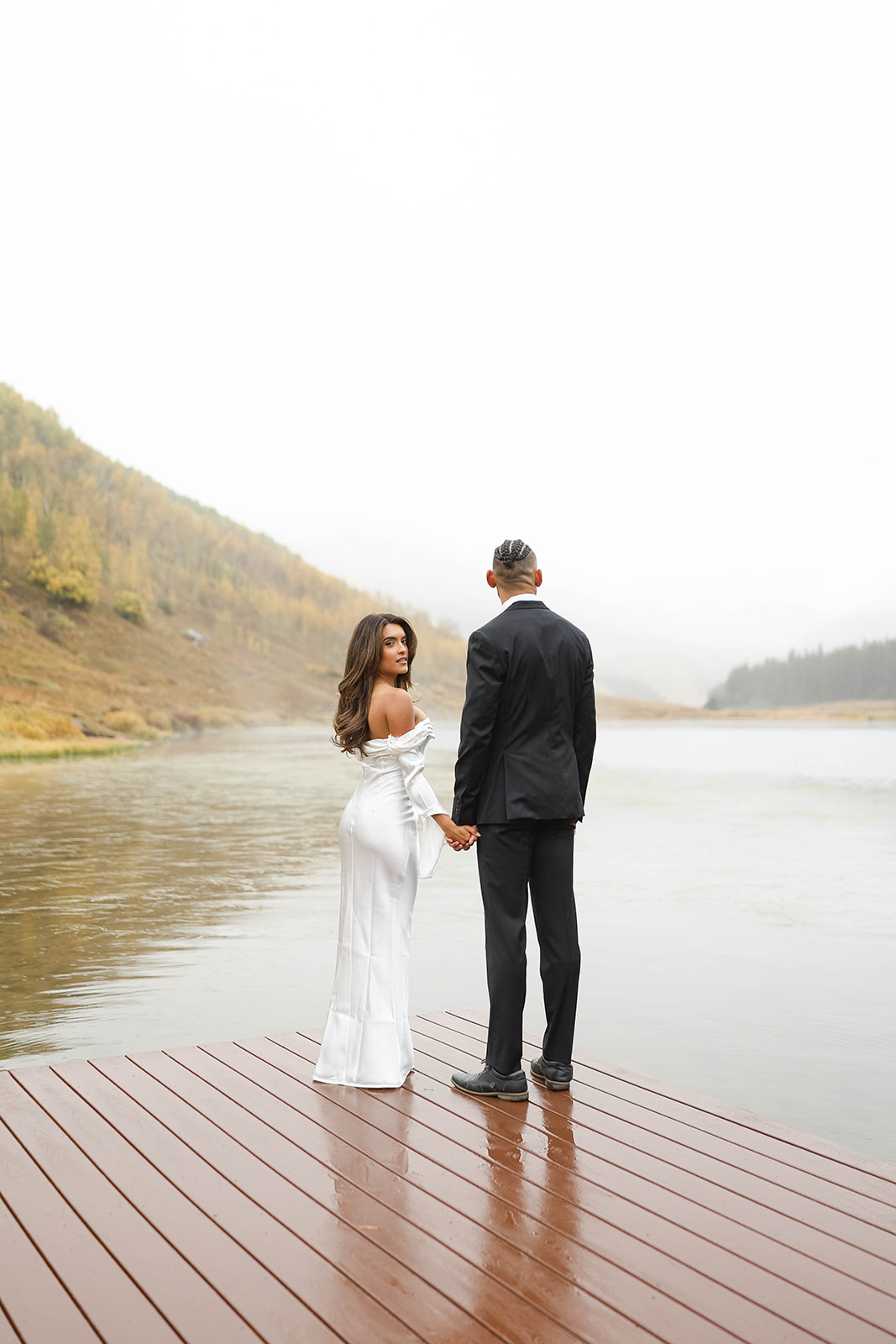 Couple holding hands on a wooden dock overlooking a calm lake, the bride glancing back toward the camera during a rainy vail elopement.