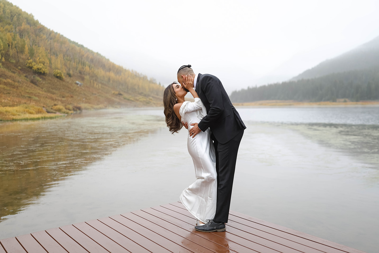A couple kissing on a dock at a mountain lake, with misty water and forested hills creating a romantic vail elopement backdrop.