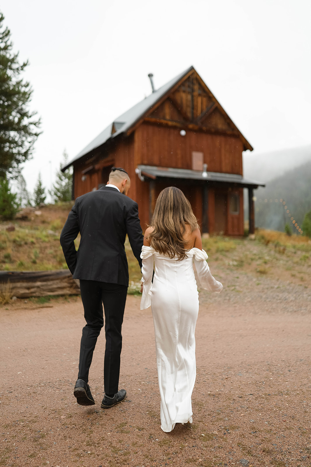 Couple walking hand in hand toward a rustic wooden cabin during a misty mountain vail elopement, with the bride in an off-the-shoulder white dress and the groom in a black suit.